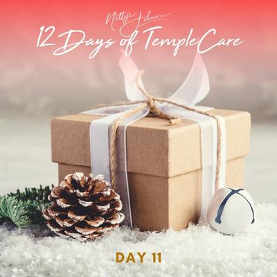 Brown gift box with white ribbon next to pine greenery, a pinecone, and a small bell on snow